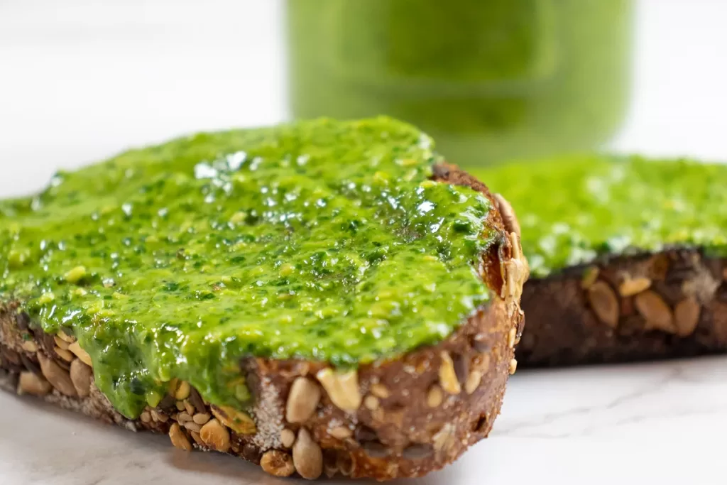 Two slices of whole-grain bread topped with bright green pesto. The bread has visible seeds and grains, and a jar of green pesto is blurred in the background.